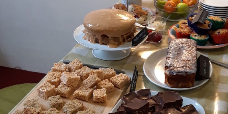 A table of food including several cakes, a plate of flapjacks and a bowl of fruit