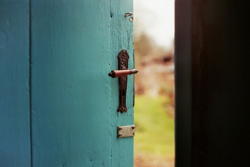 A close-up of a wooden door with a prominent handle, slightly ajar to reveal the countryside. The image signifies transition