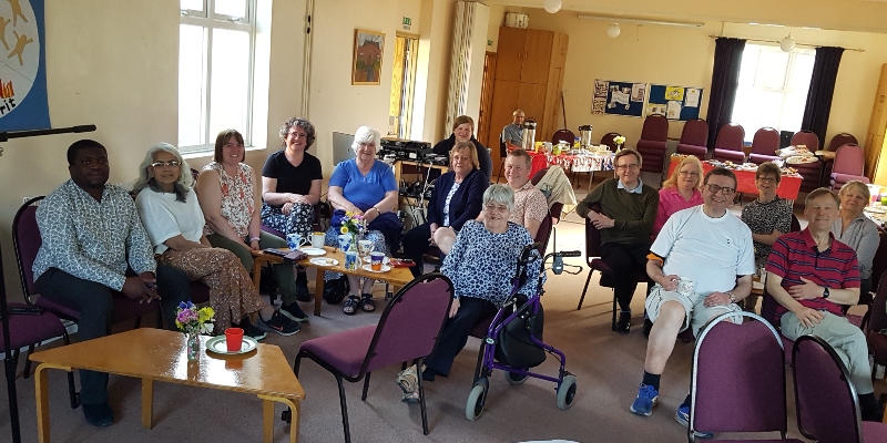16 people at the Hemel small church hub sit on chairs facing the camera and around a couple of small tables