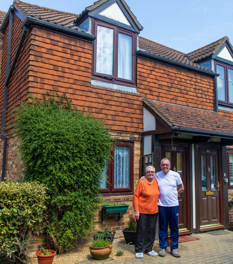 A retired man and woman stand in front of a house