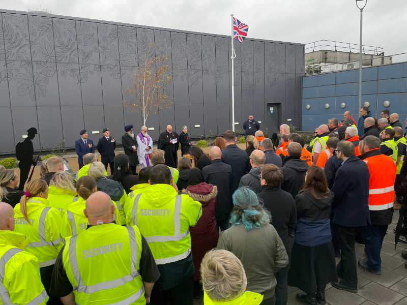 Remembrance service at East Midlands Airport showing around 30-40 members of staff, some wearing high visibility jackets, listening to clergy
