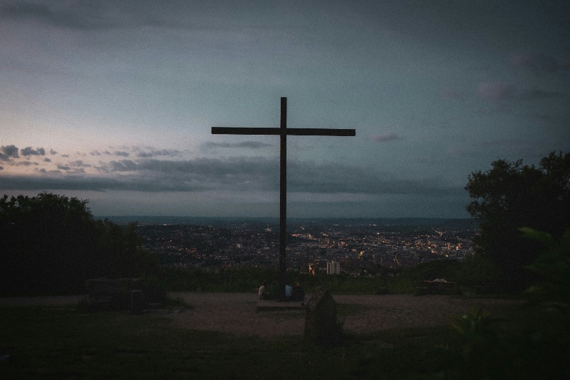 A large cross silhouetted against a twilight sky overlooks a distant city with scattered lights. Trees frame the scene