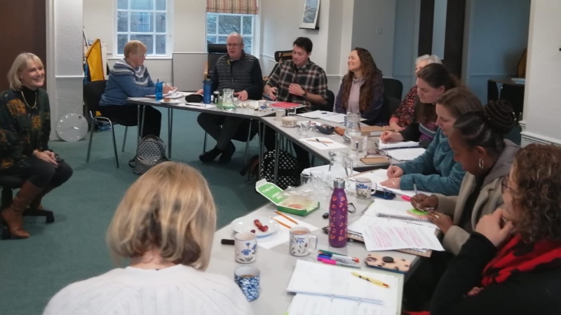 A previous Paths of Peace Course, showing A group of ten people sit around a U-shaped table in a meeting room, engaging in discussion. Papers, notebooks, and mugs are on the table. Mediation trainer and Baptist minister Joanna Williams sits at the front