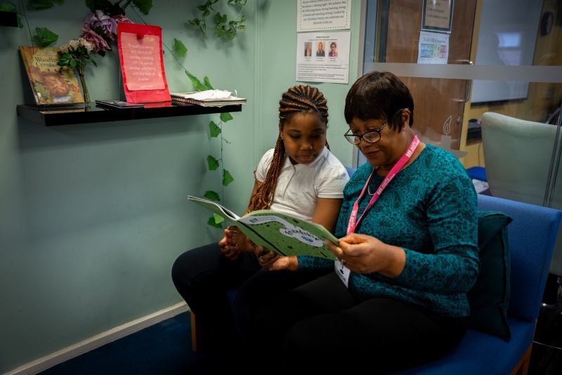 A woman in glasses and a young girl read a book together on a blue couch in a cozy room with books and decor