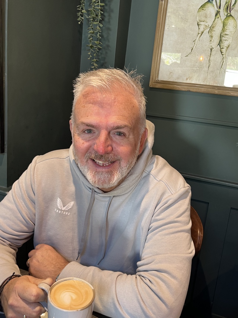 Mark Elder, smiling in a cafe with one hand holding a cup of coffee
