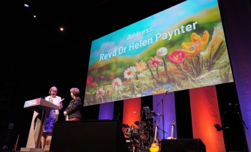 Lynn Green and Helen Paynter stand on a stage with a large screen displaying "Address: Revd Dr Helen Paynter" and vibrant flower art. Musical instruments are visible below the screen.
