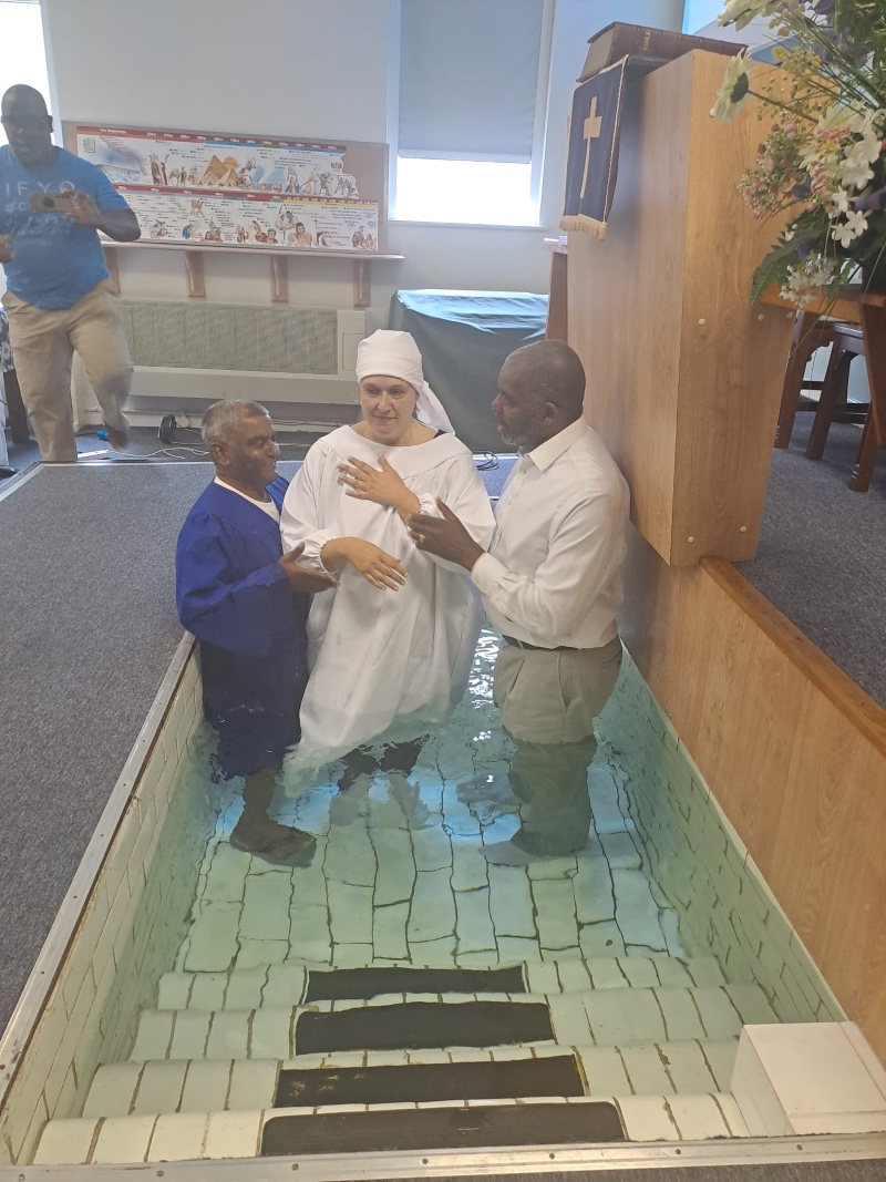A baptism is taking place in a small indoor pool with three people. One person in white is being baptized by two others in white and blue robes.