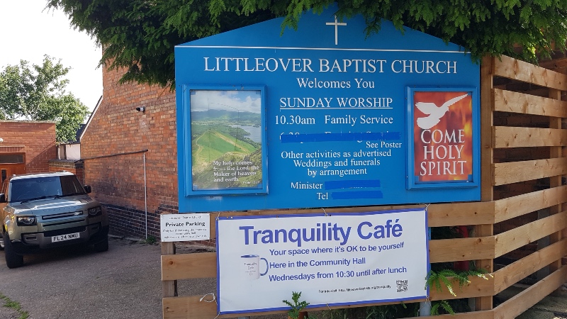 A large blue sign for Littleover Baptist Church displays service information and features two smaller poster display cases. Below it, a white banner advertises "Tranquility Café" held on Wednesdays in the Community Hall. A modern grey SUV is parked in a driveway to the left of the sign.