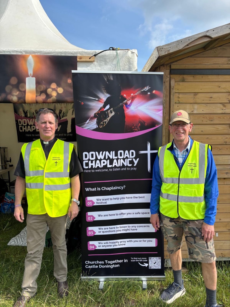 Two men in high-visibility "Festival Chaplain" vests standing outdoors next to a "Download Chaplaincy" roll-up banner outside a marquee.