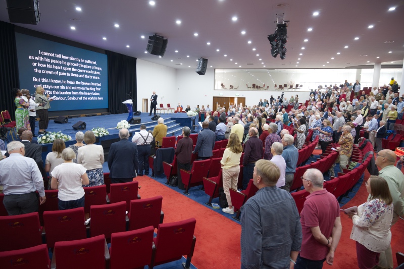 A large congregation standing in a modern auditorium during a worship service, facing a stage with song lyrics projected on a large screen