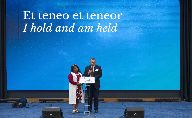 Alex and Dorcas Afriyie, standing at a Trinity lectern on stage in front of a large blue screen displaying the words "Et teneo et teneor" and "I hold and am held".
