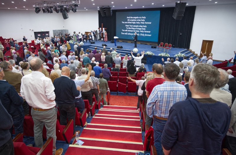 A large congregation standing in a modern auditorium during a worship service, facing a stage with song lyrics projected on a large screen