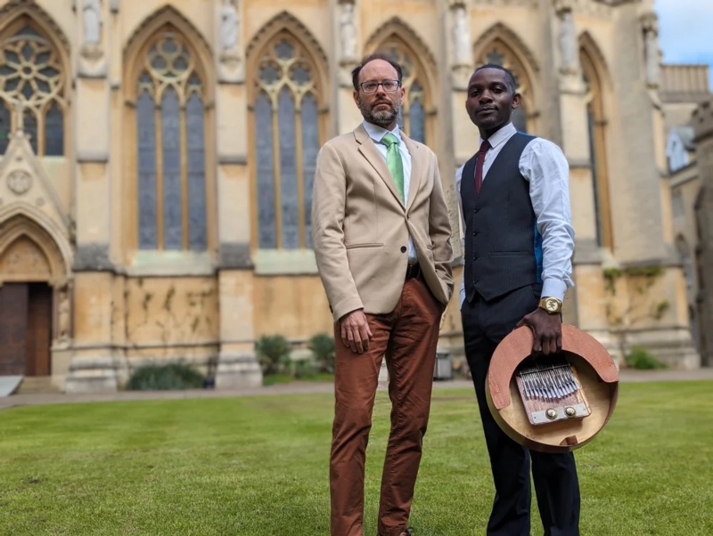 Two smartly dressed men standing outdoors on a lawn in front of a historic gothic church building, with one holding a thumb piano instrument.